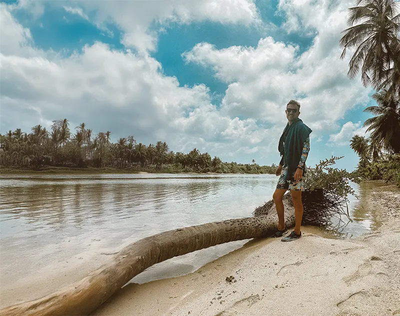 Mann am tropischen Strand neben Palmen und Wasser unter blauem Himmel im Hintergrund.