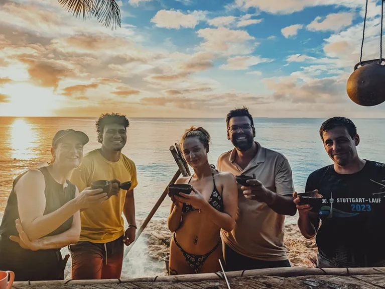 Gruppe genießt tropischen Sonnenuntergang am Strand, fröhlich mit Getränken in der Hand.