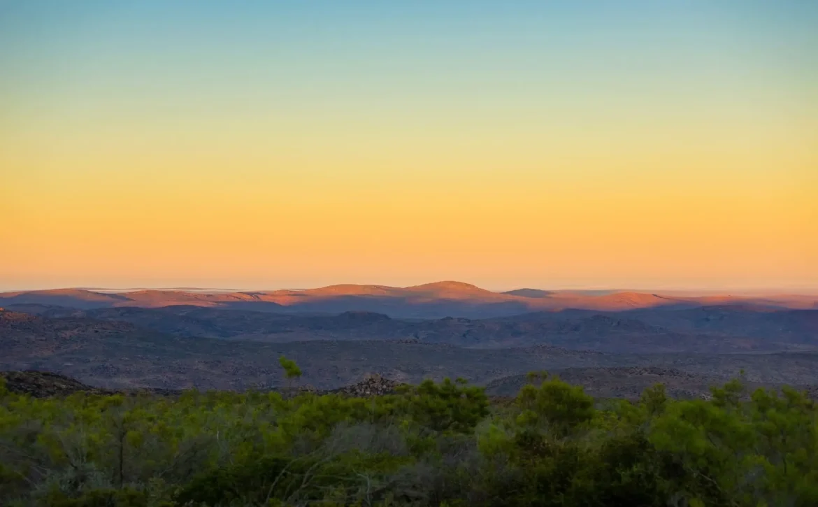 Sonnenuntergang über kargen Hügeln, goldenes Licht am Horizont, bewölkter Himmel, weite Landschaft, friedliche Natur.