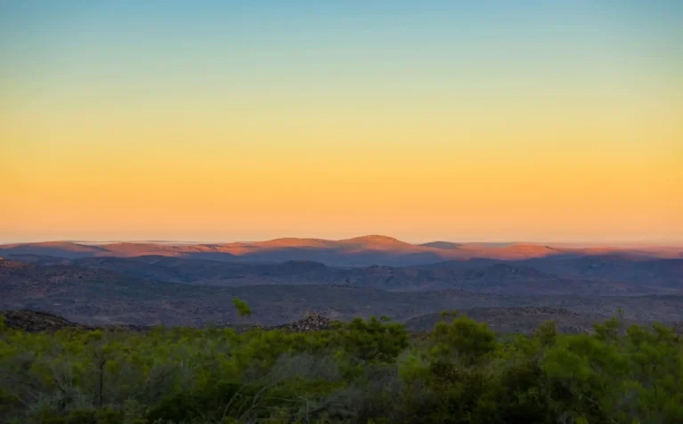 Sonnenuntergang über kargen Hügeln, goldenes Licht am Horizont, bewölkter Himmel, weite Landschaft, friedliche Natur.