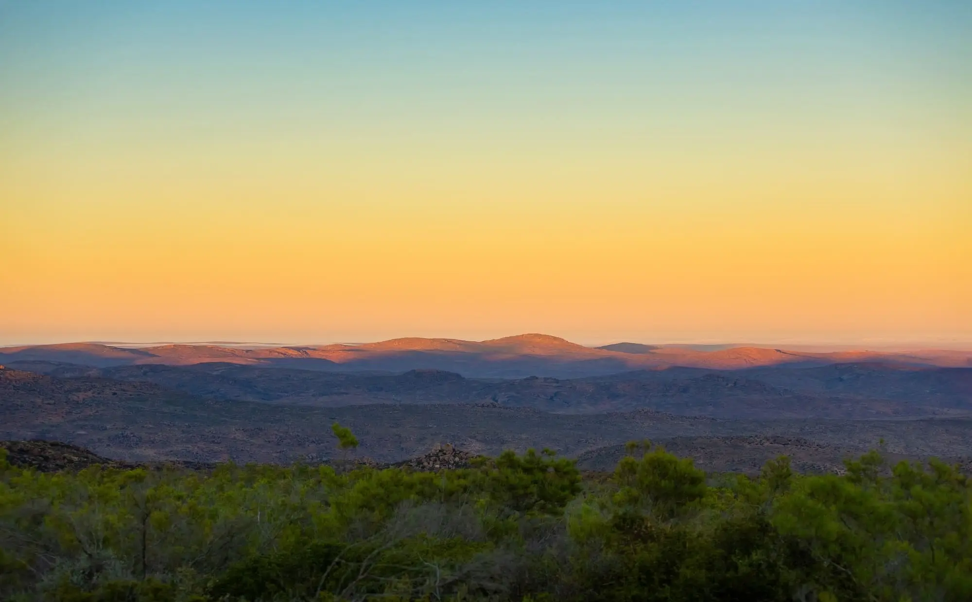 Sonnenuntergang über kargen Hügeln, goldenes Licht am Horizont, bewölkter Himmel, weite Landschaft, friedliche Natur.