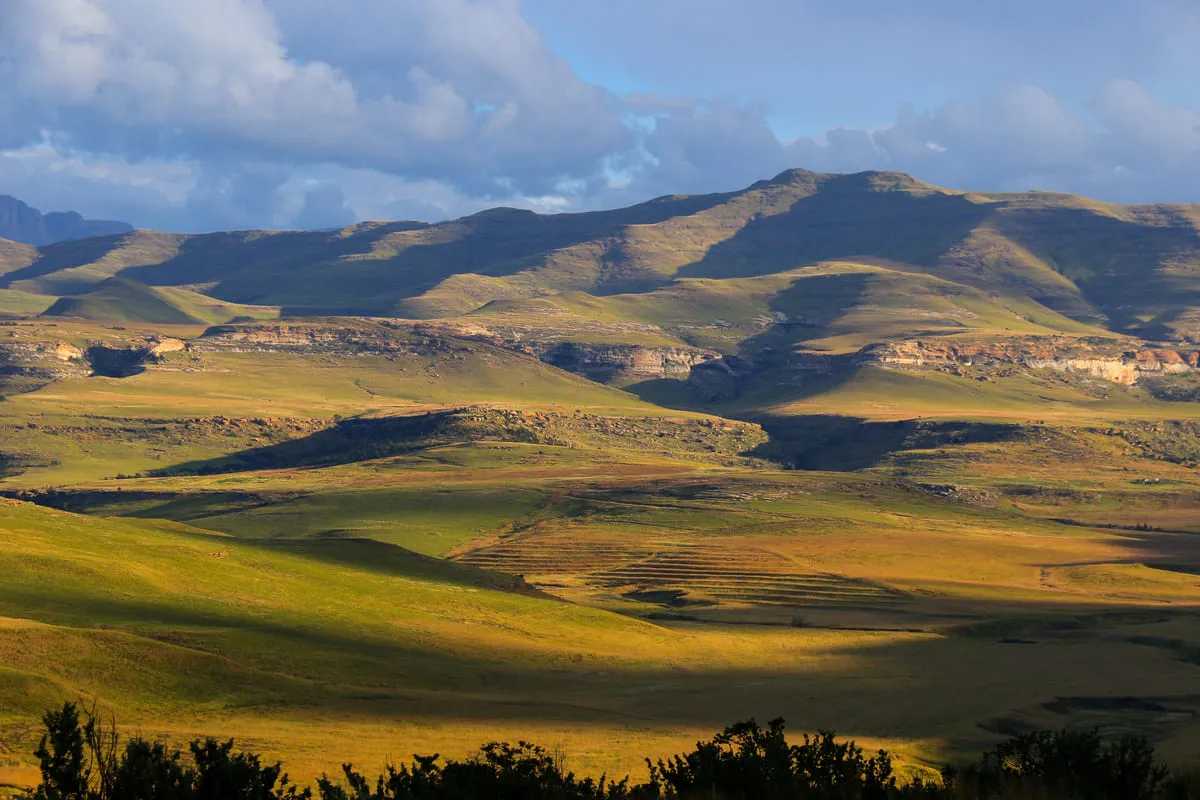 Sanfte Hügel und weite Graslandschaft unter dramatischem Himmel mit Wolken und Sonnenlichtspiel.