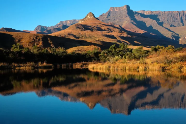Berglandschaft mit klarer Spiegelung im See, sonnendurchflutete Hügel und blauer Himmel.