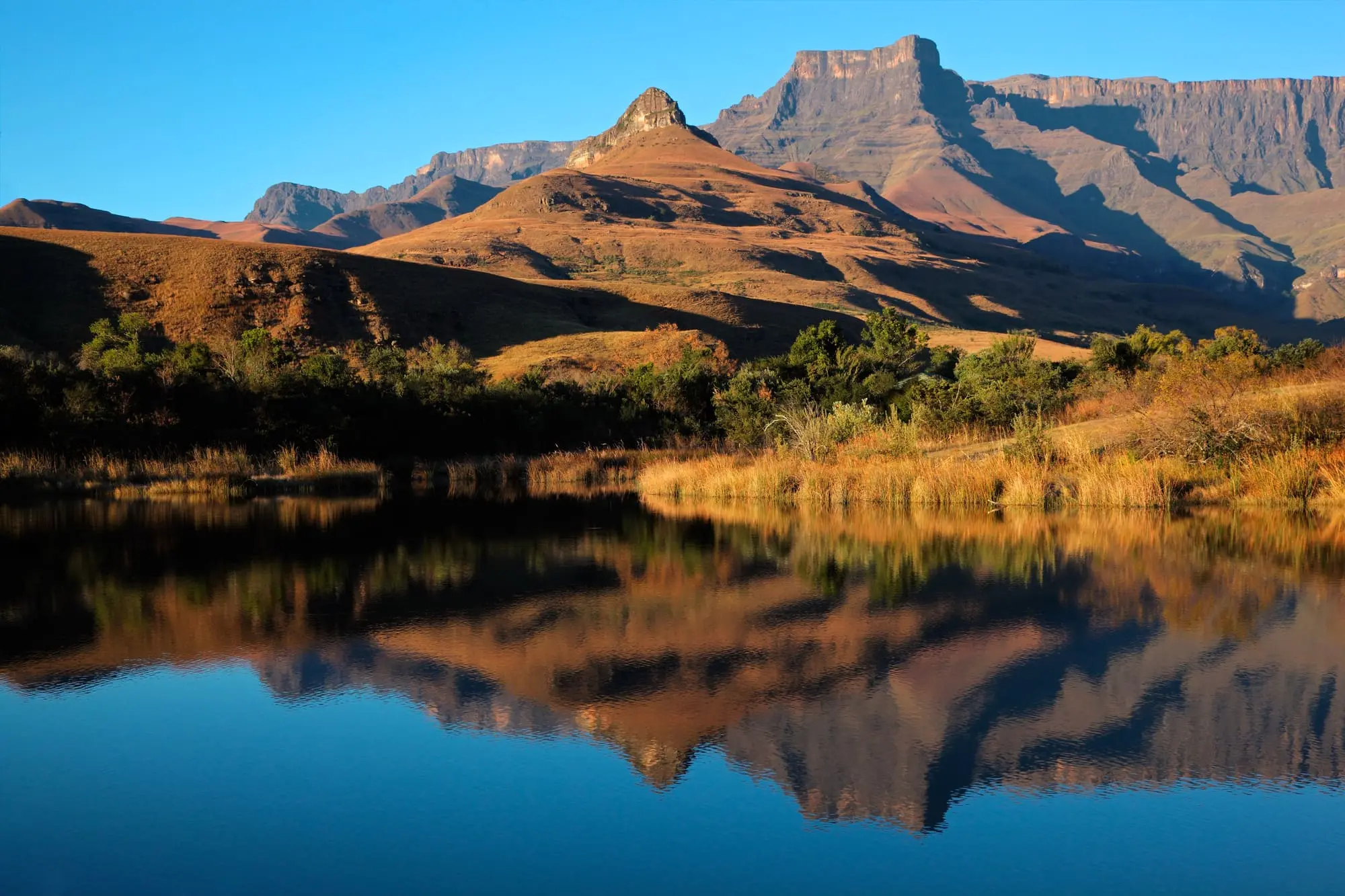 Berglandschaft mit klarer Spiegelung im See, sonnendurchflutete Hügel und blauer Himmel.