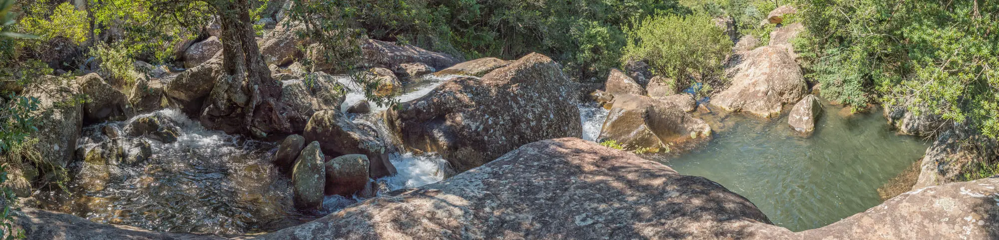 Natürliche Felsformationen und kleiner Wasserfall in dicht bewaldeter Landschaft mit klarem Wasser.