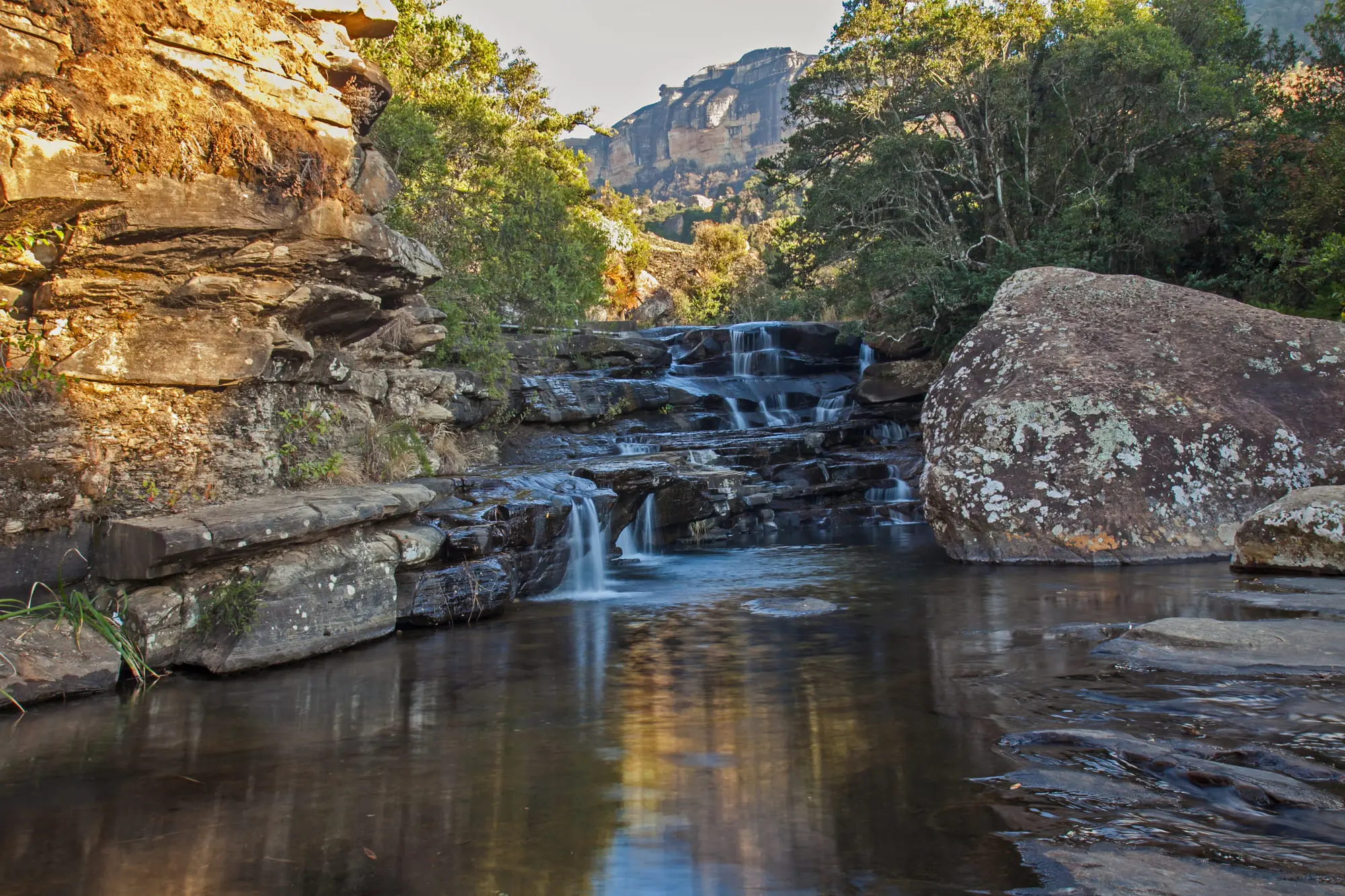 Kaskadierende Wasserfälle in ruhiger Naturkulisse, umgeben von Felsen und üppigem Grün unter strahlendem Himmel.