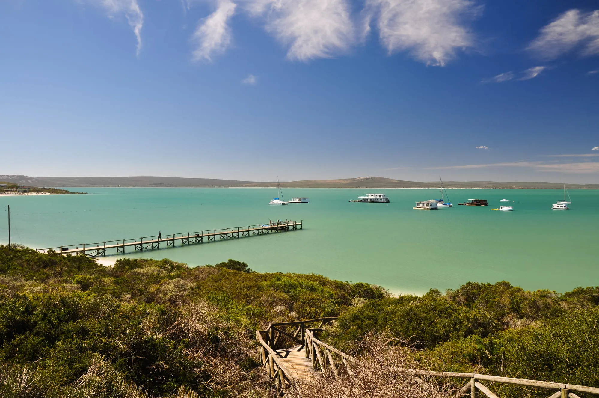 Holzsteg führt durch Dünen zu türkisfarbenem Meer mit Segelbooten, unter klarem Himmel – idealer Küstenpanorama.