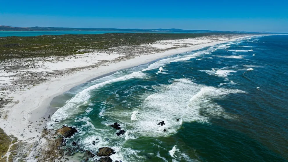 Weitläufiger Sandstrand mit türkisfarbenem Meer und weißen Wellen an der Küste unter blauem Himmel.