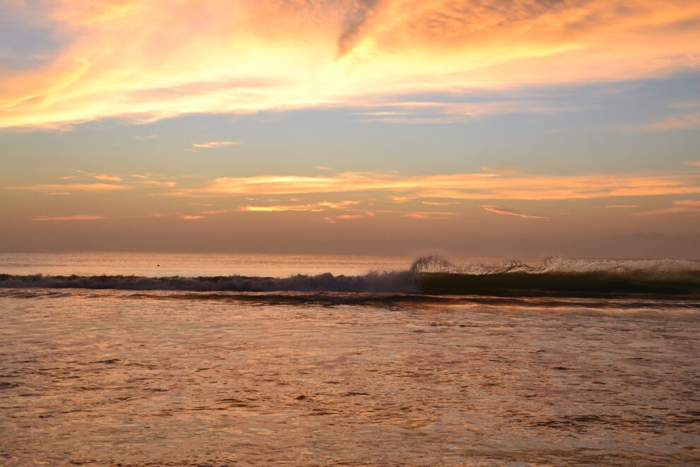 Abendrot über der Bucht von Jimbaran auf Bali, eine Welle bricht im warmen Licht des Sonnenuntergangs