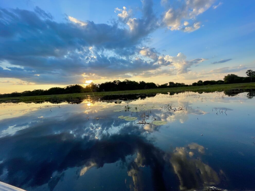 Der Thamalakane-Fluss in Maun am Abend, Wolken und die dunkle Baumreihe spiegeln sich im Wasser