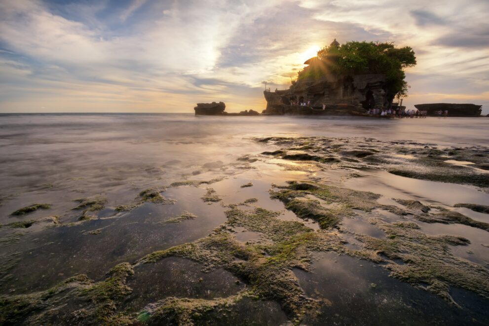 Sonnenuntergang hinter dem Felsentempel Pura Tanah Lot auf Bali, im Vordergrund das moosige Felsplateau bei Ebbe