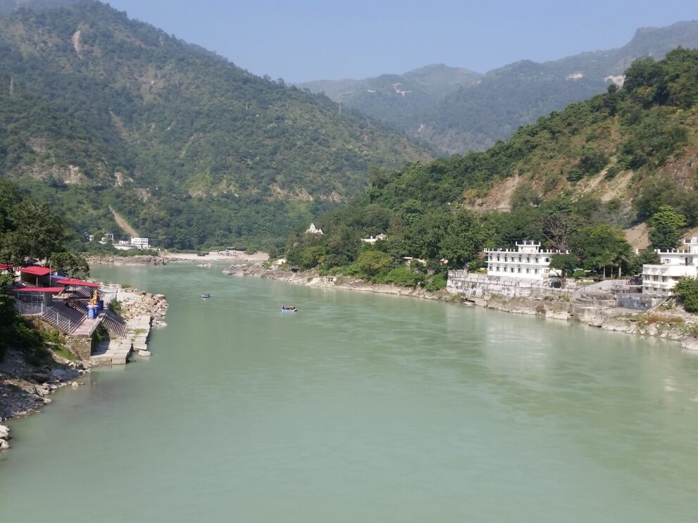 Der Ganges in Rishikesh zwischen bewaldeten Hängen der Himalaya-Ausläufer, am Ufer Ashrams und Ghats