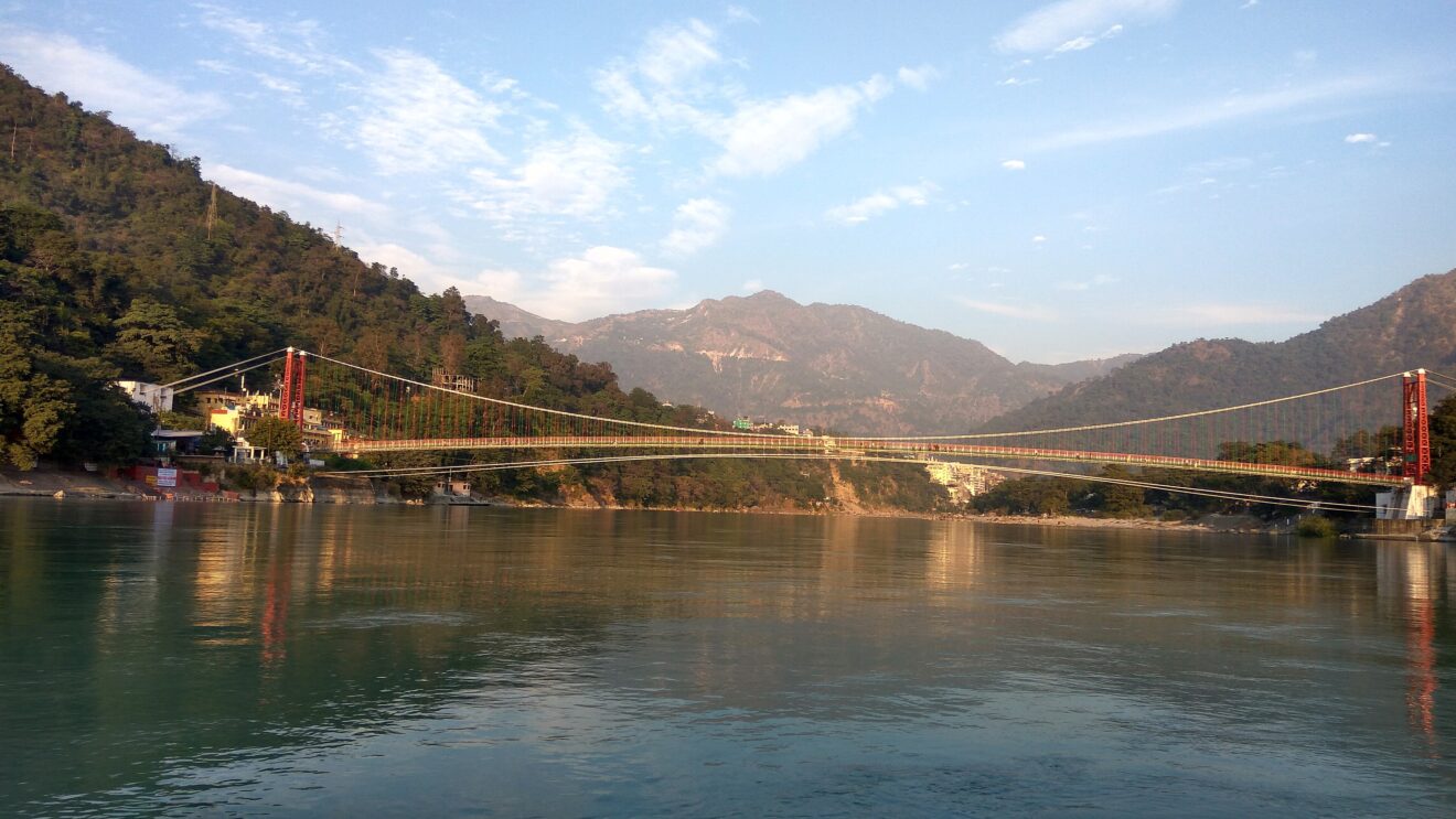 Die Hängebrücke Lakshman Jhula über den Ganges in Rishikesh, vom Boot aus gesehen, dahinter die Berge