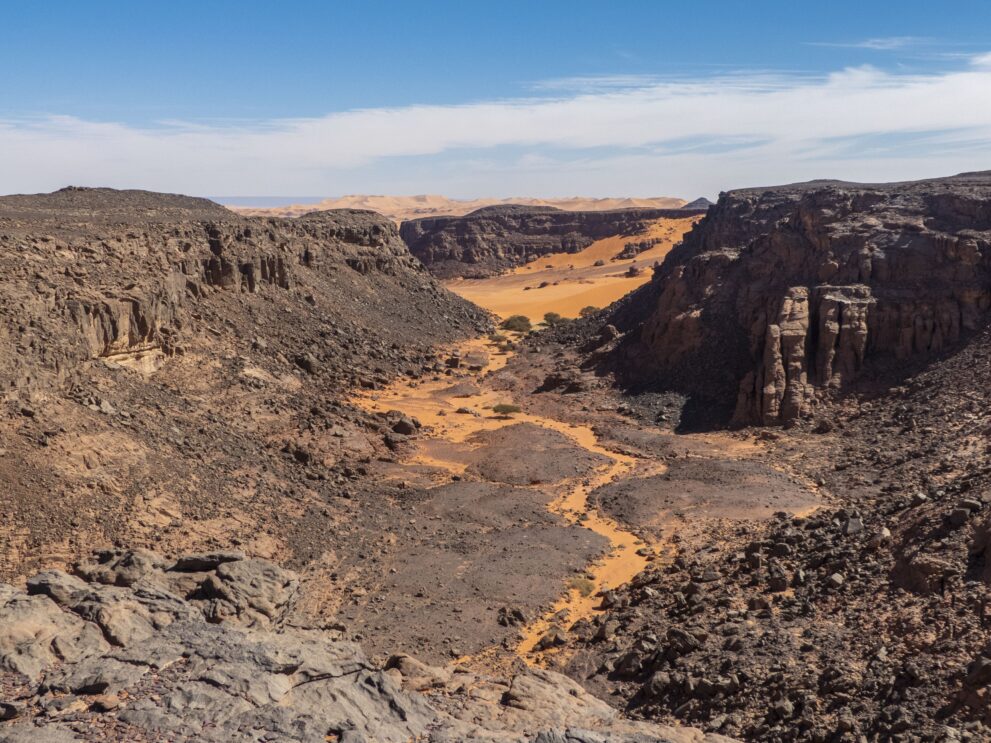Trockenes Wadi mit Sandband zwischen dunklen Felshängen bei Bouhadian im Roten Tadrart