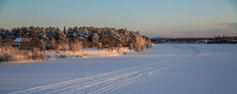 Zugefrorener Fluss Ivalojoki mit Schneemobilspuren und verschneitem Ufer bei Ivalo im Winterlicht