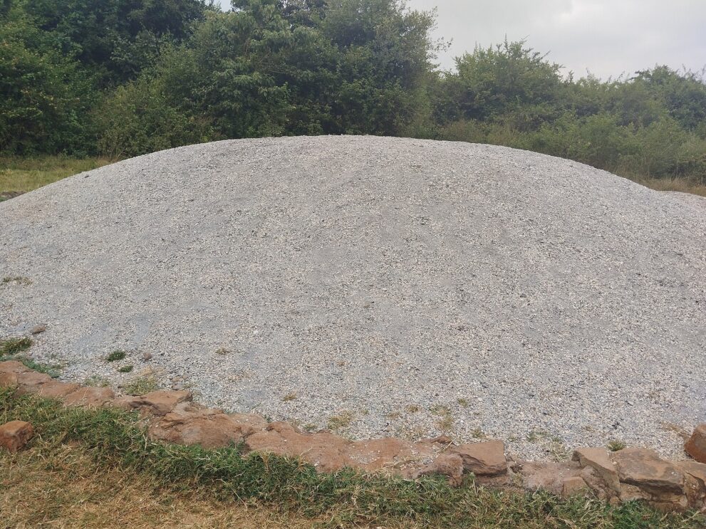Flacher Hügel aus grauer Asche in einer Einfassung aus rötlichen Steinen, dahinter dichtes Buschwerk