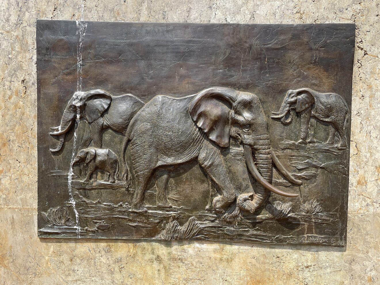 Bronzerelief einer Elefantenfamilie mit Jungtier in flachem Wasser, eingelassen in eine helle Steinwand, signiert 1989