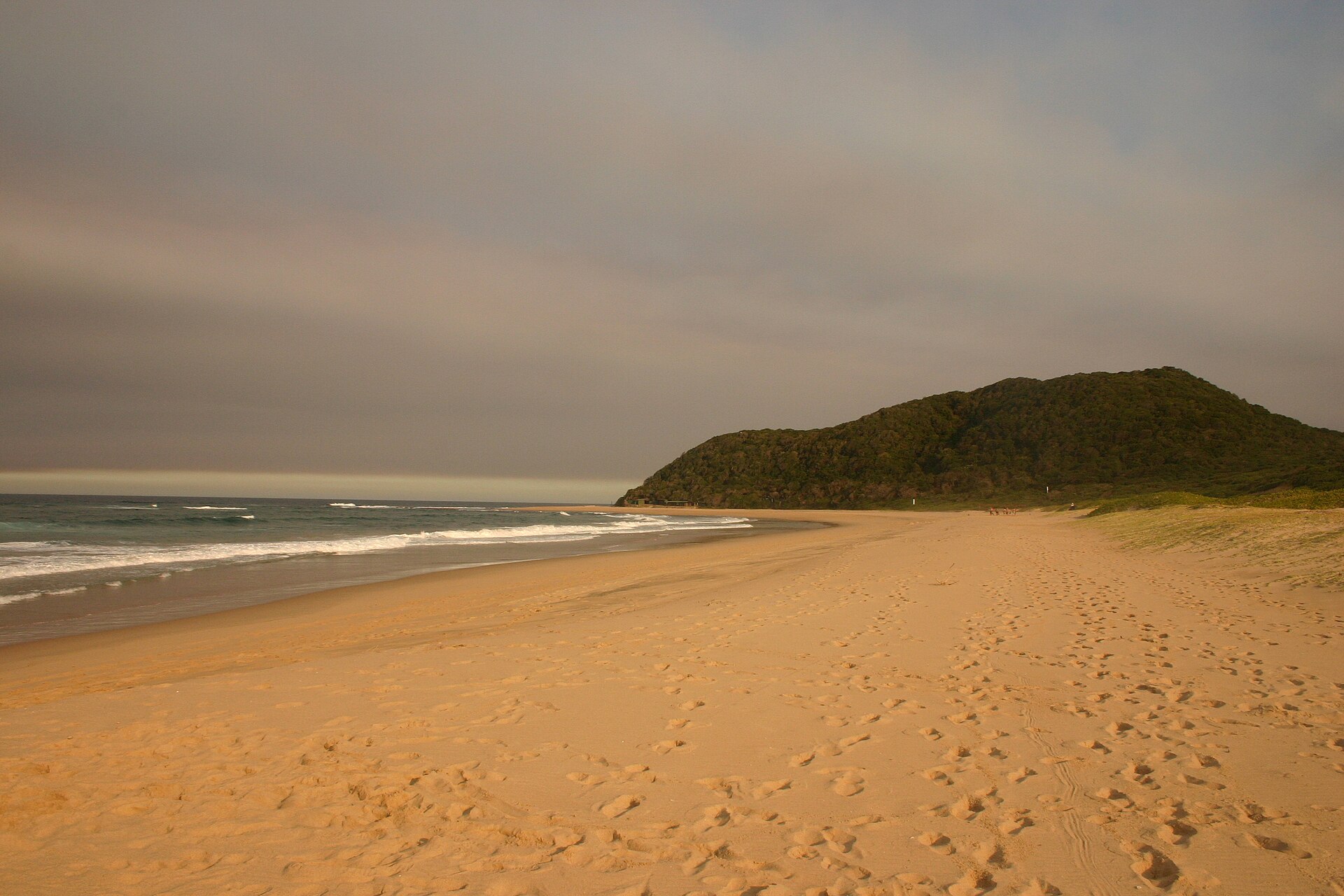 Breiter, leerer Sandstrand mit Fußspuren im Abendlicht, rechts ein bewaldeter Dünenhügel am Meer