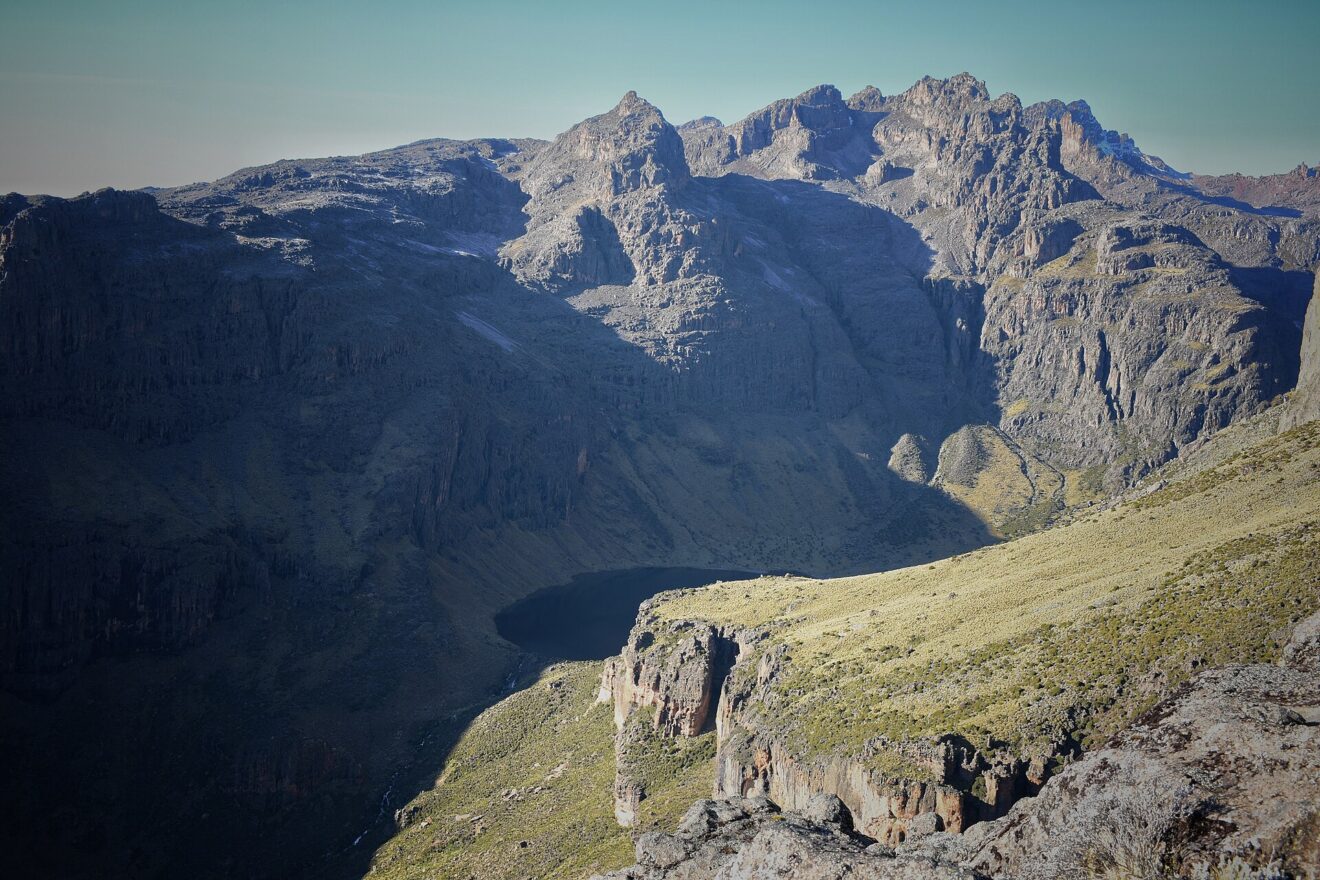 Blick von oben in einen tiefen Felskessel: unten ein dunkler See, dahinter zackige Gipfel und steile Wände im Morgenlicht.