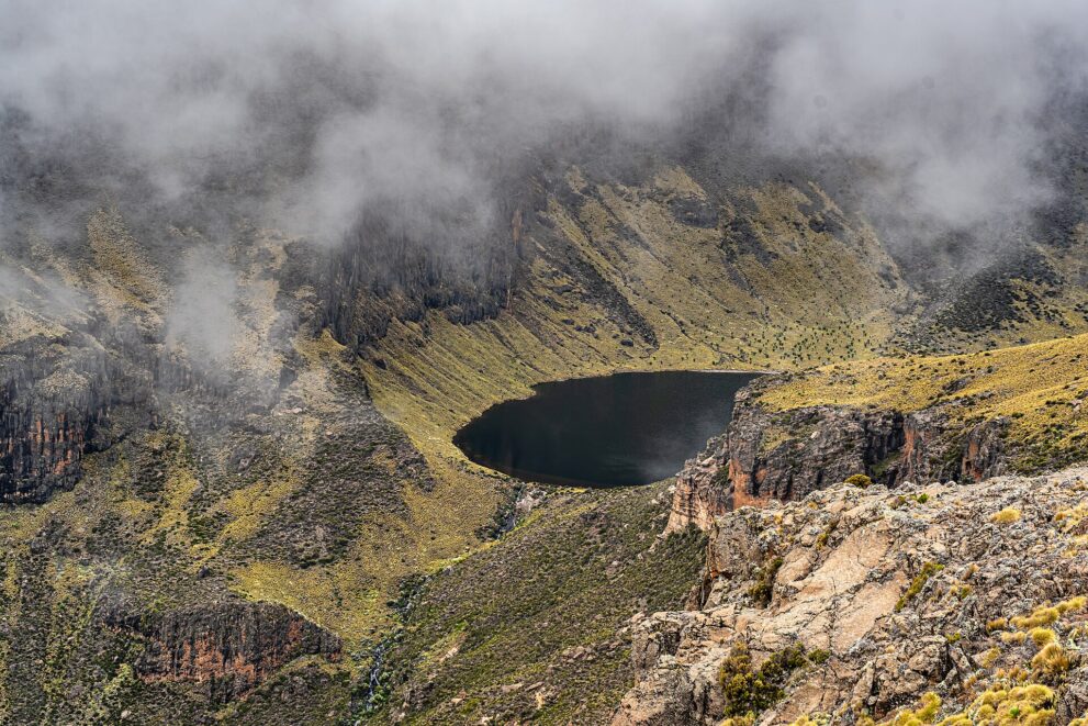 Dunkler See in einem grünen Felskessel, über den Wänden hängen tiefe Wolken, vorn heller Fels mit Grasbüscheln.