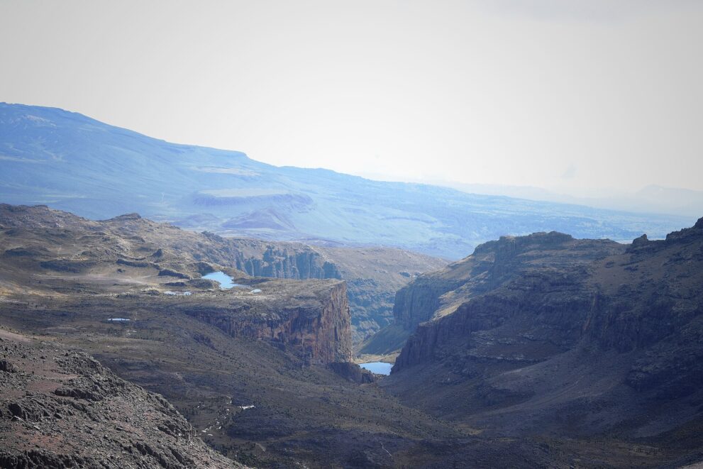 Weiter Blick von einem Gipfel hinab: auf einer Hochfläche kleine blaue Seen, darunter eine Felswand und tief im Tal ein weiterer See.