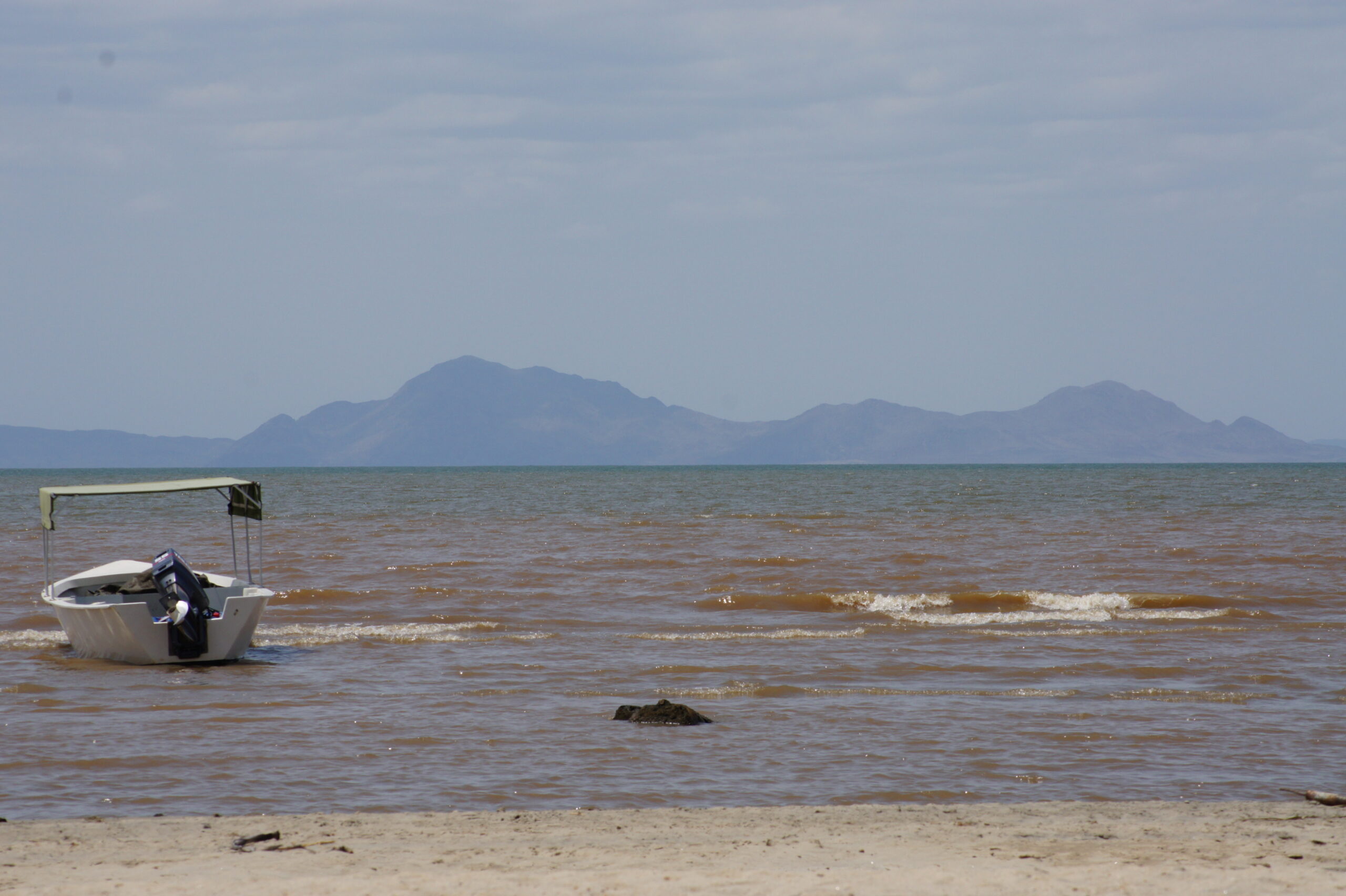 Ein kleines Motorboot mit Sonnendach liegt im flachen, braunen Wasser, kurze Wellen laufen ans Ufer, dahinter Berge im Dunst