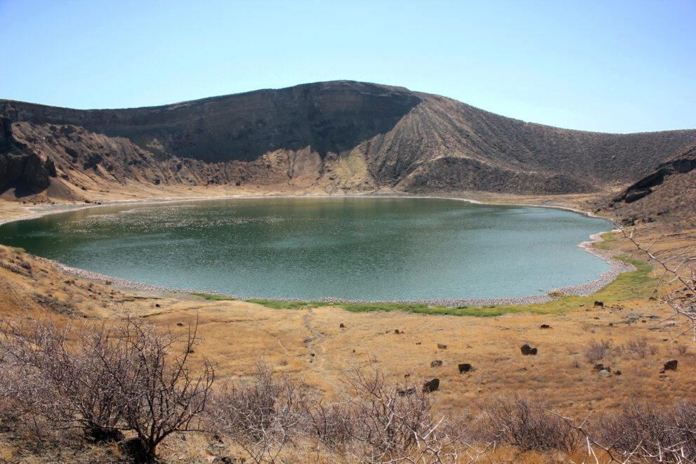 Grüner Kratersee in einem steilen, kahlen Vulkankrater, am Ufer ein heller Saum, vorn trockenes Gras und dorniges Gebüsch, Central Island im Lake Turkana