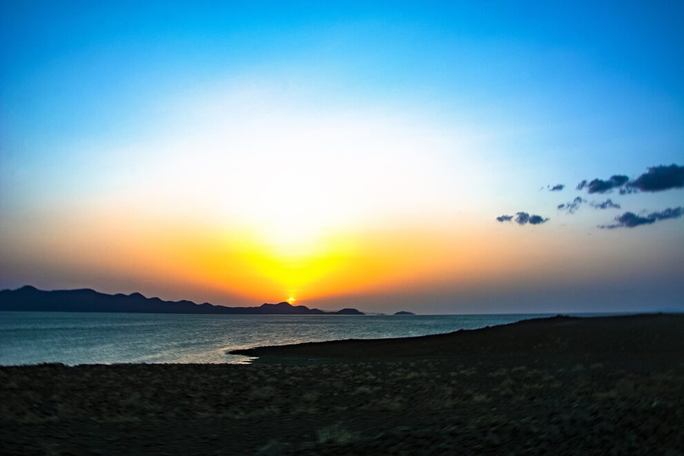 Sonne geht hinter Bergen am Turkana-See unter, vorne dunkles Steinufer