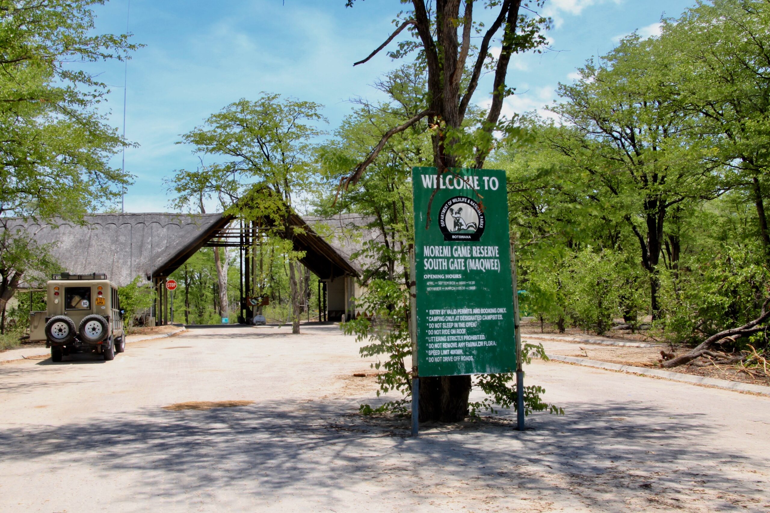 Das South Gate des Moremi-Game-Reserve mit Reetdach über der Zufahrt, davor ein Geländewagen und das grüne Schild der Wildtierbehörde mit den Öffnungszeiten