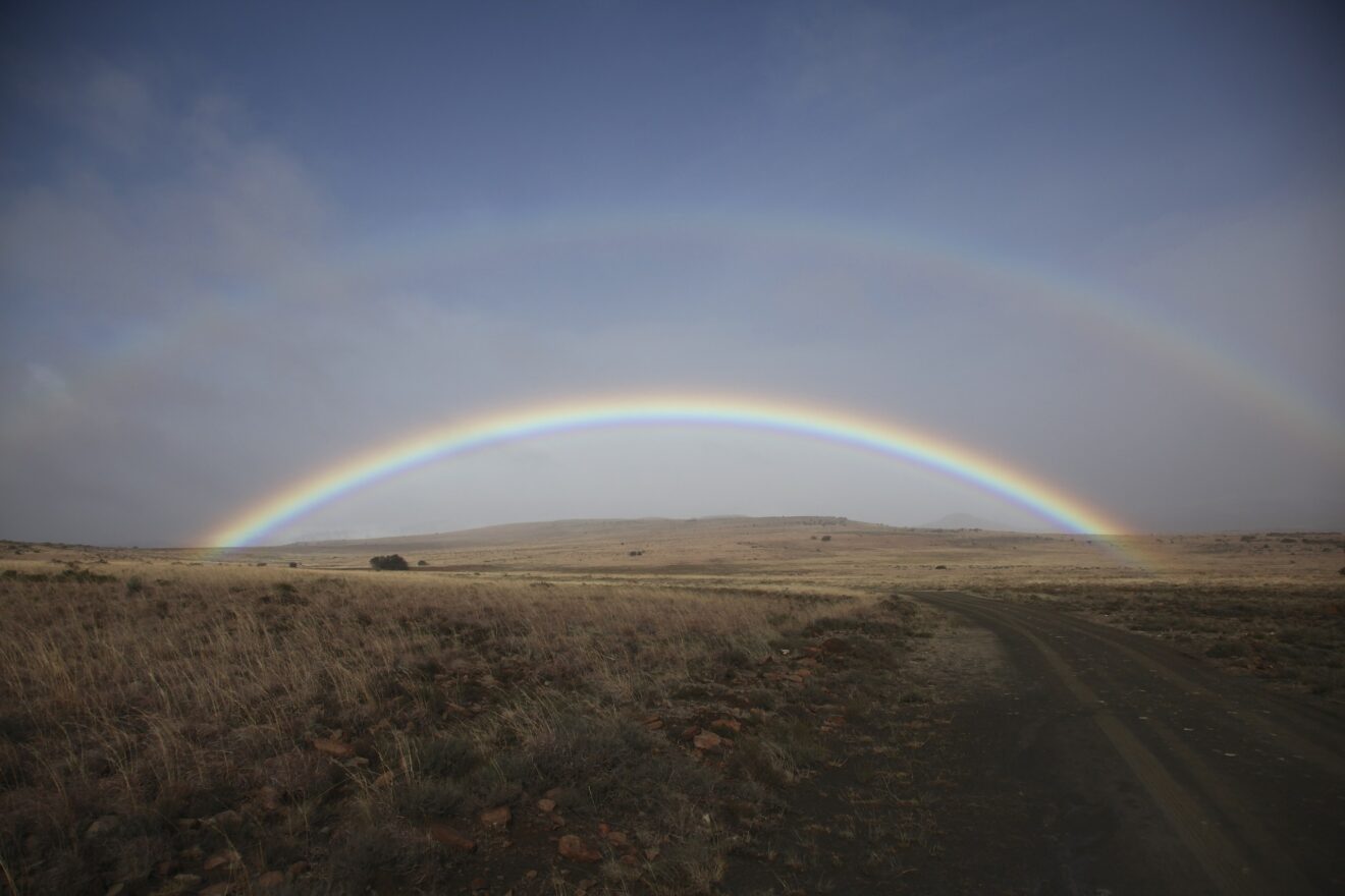 Doppelter Regenbogen über einer weiten, trockenen Hochebene mit Schotterstraße unter grauem Himmel