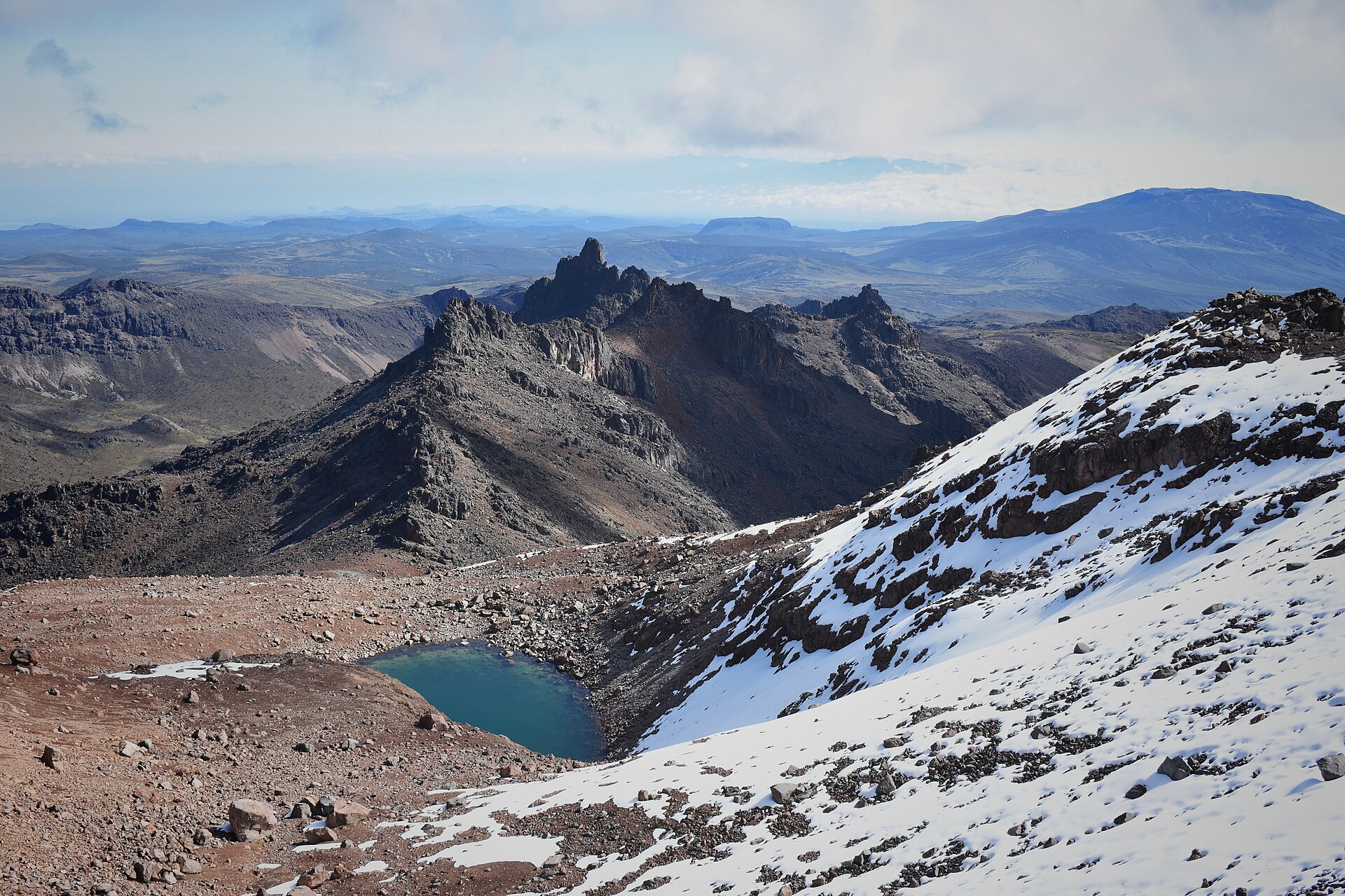 Verschneiter Hang, darunter ein kleiner türkiser Bergsee, dahinter ein zerklüfteter Grat und weites Hochland