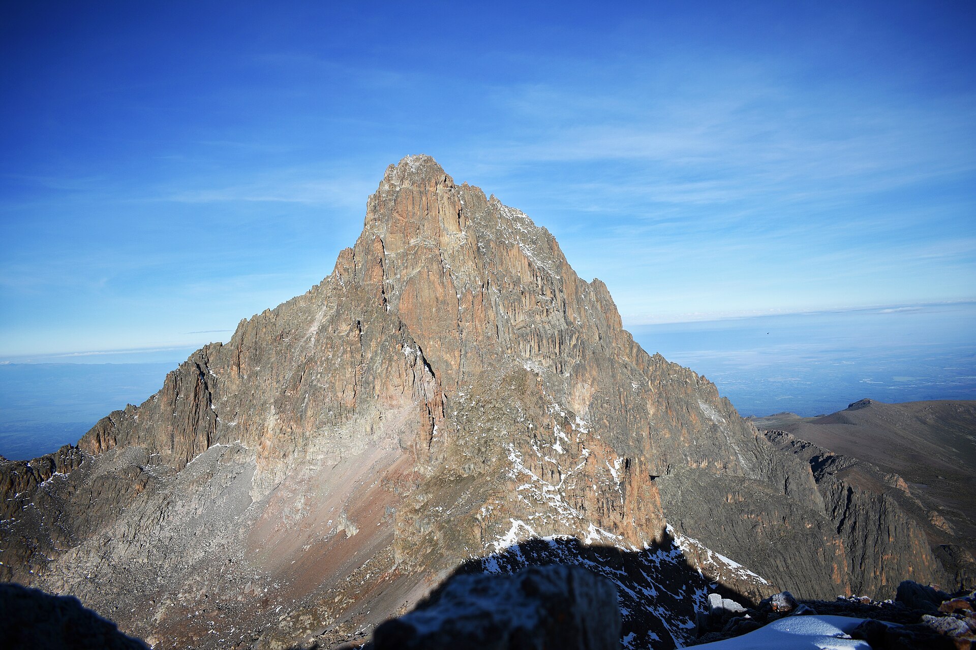 Mächtiger rötlicher Felsturm mit Schneeresten vor blauem Himmel, rechts hinten das Hochland