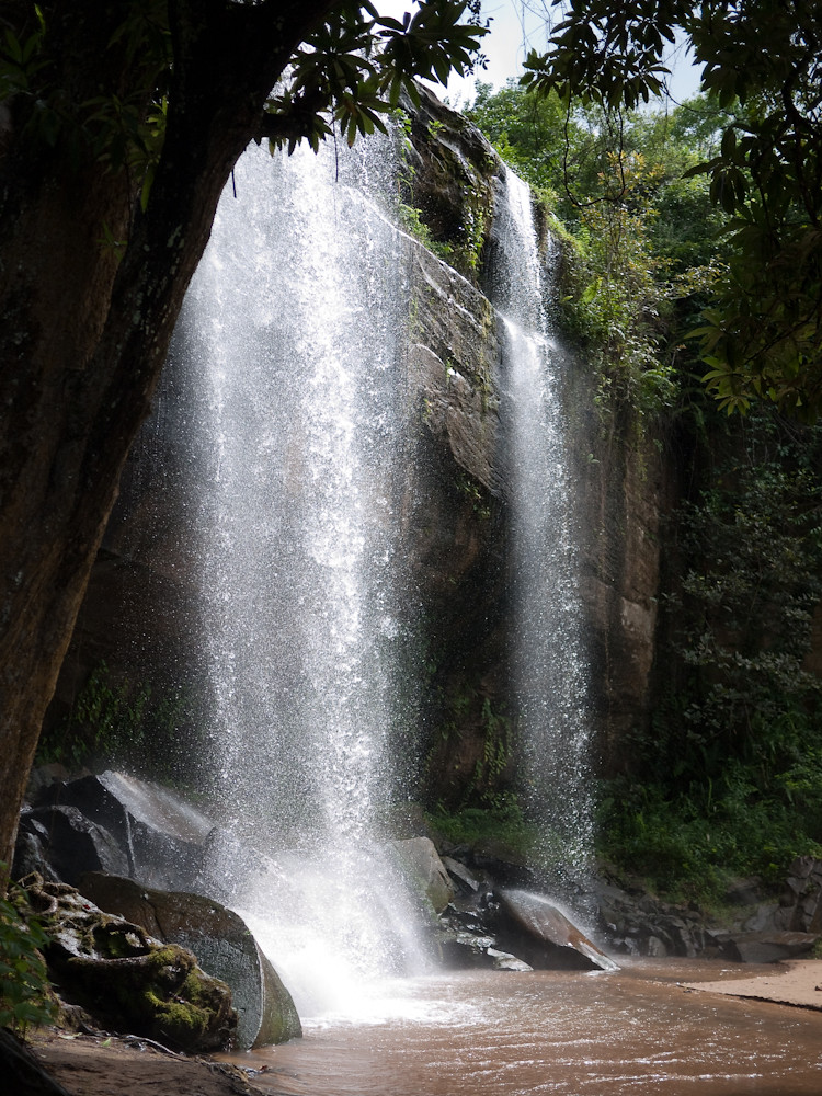 Zwei kräftige Wasserschleier fallen über eine dunkle Felswand in ein Becken mit Felsblöcken, links ein Baumstamm