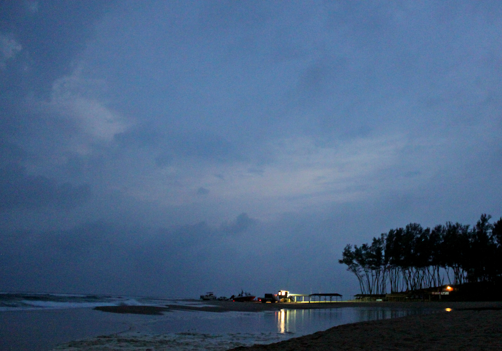 Blaue Morgendämmerung an einem Strand, Boote auf Anhängern an der Startstelle, rechts Kasuarinen und ein beleuchteter Unterstand