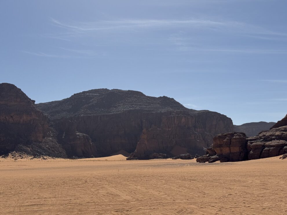 Dunkle Felswände und ein weites Sandtal im Roten Tadrart unter blauem Himmel, Südosten Algeriens