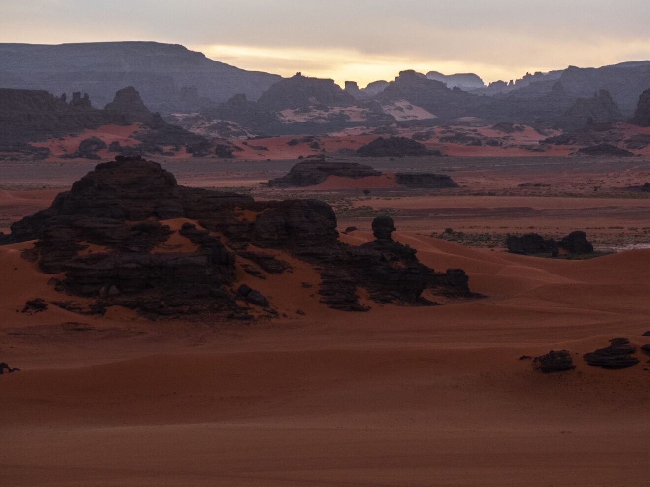 Abendlicht über den dunklen Felstürmen und roten Sandflächen von Tin Merzouga im Roten Tadrart
