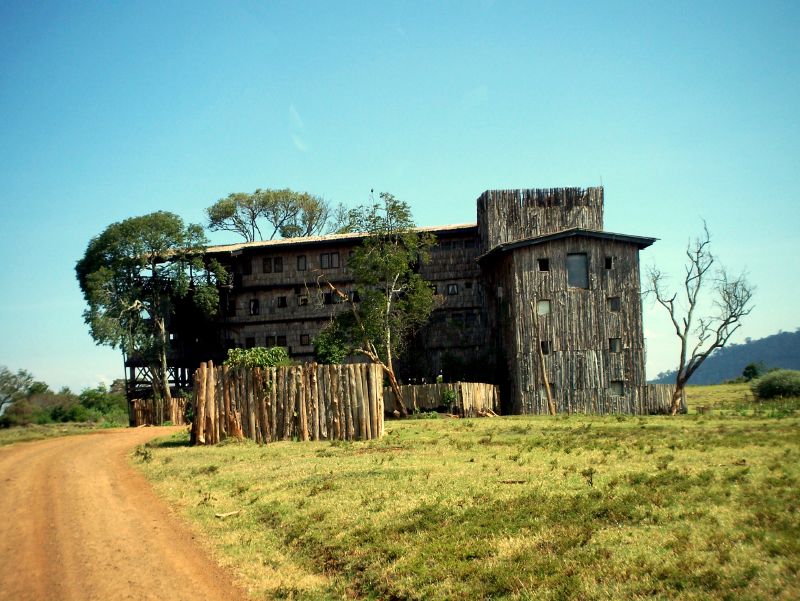Hoher Holzbau mit Turm hinter einer Palisade aus Stämmen, davor eine rote Erdpiste und Wiese