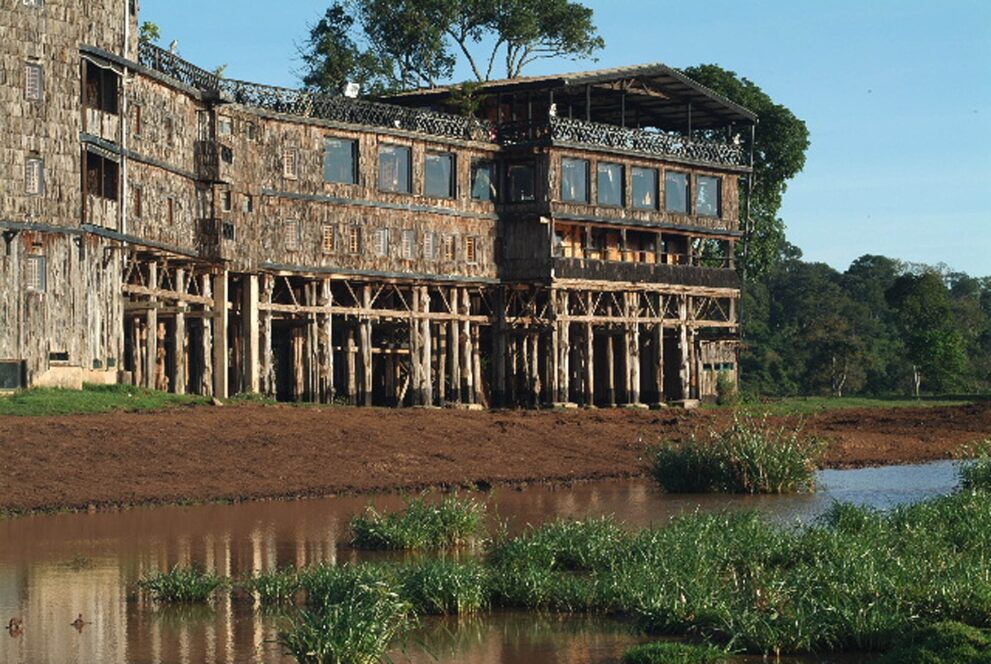 Mehrstöckiger Holzbau mit Schindelfassade auf Holzstelzen, davor ein Wasserloch mit Gras, dahinter Wald