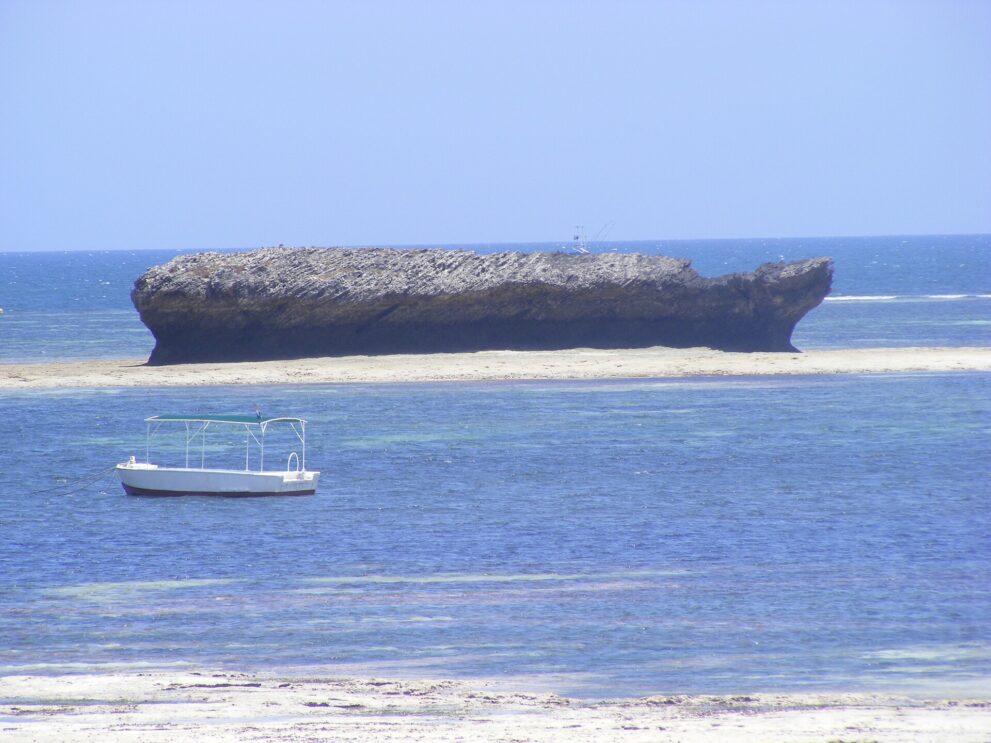 Ein weißes Ausflugsboot mit Sonnendach liegt vor einem langen, flachen Felsen, davor eine helle Sandbank im blauen Wasser