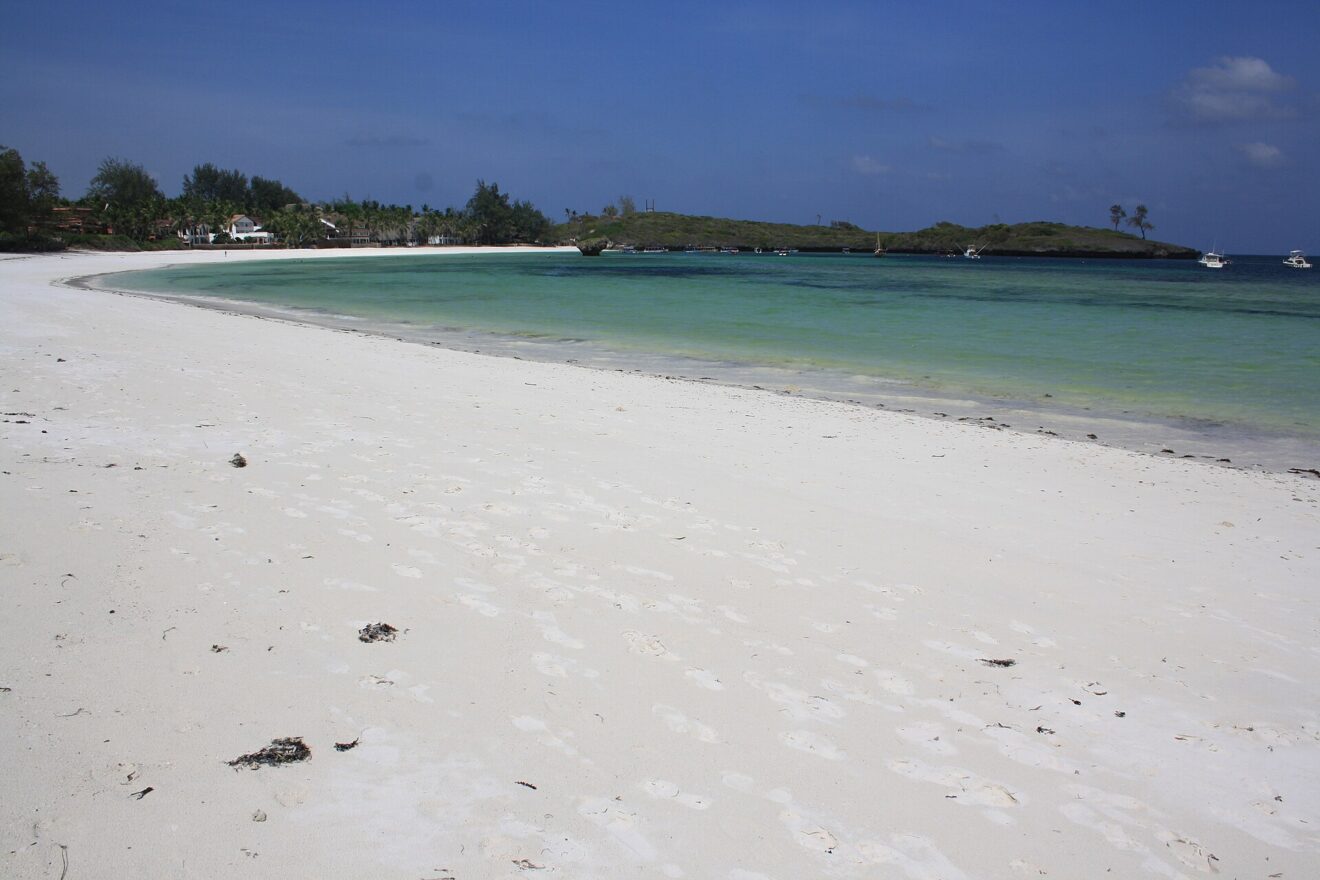 Weißer Sandstrand in weitem Bogen, türkises Wasser, dahinter eine bewachsene Landzunge und kleine Boote