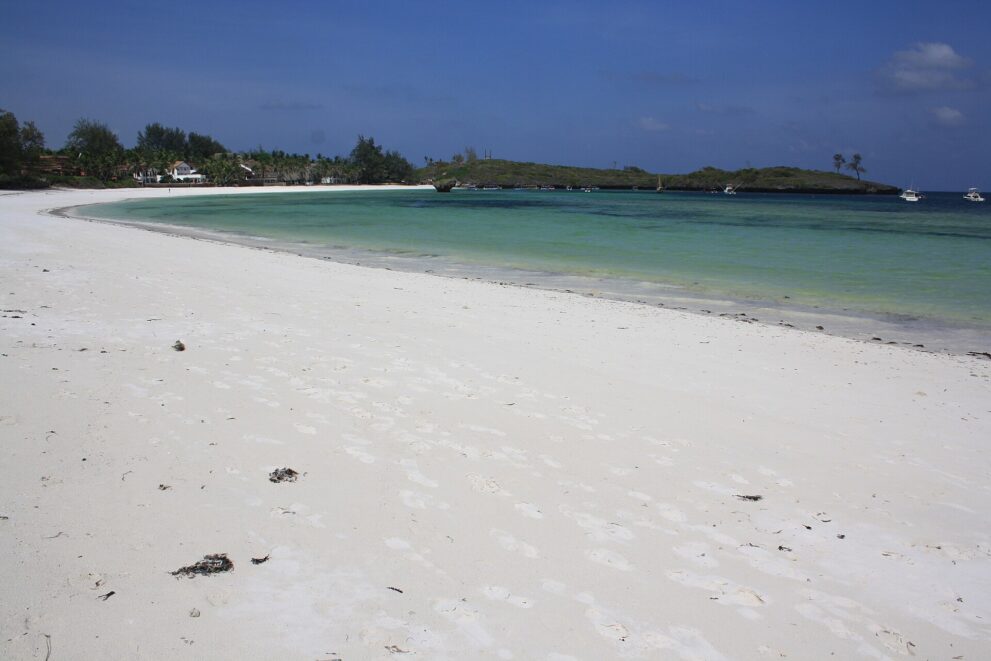 Weißer Sandstrand in weitem Bogen, türkises Wasser, dahinter eine bewachsene Landzunge und kleine Boote