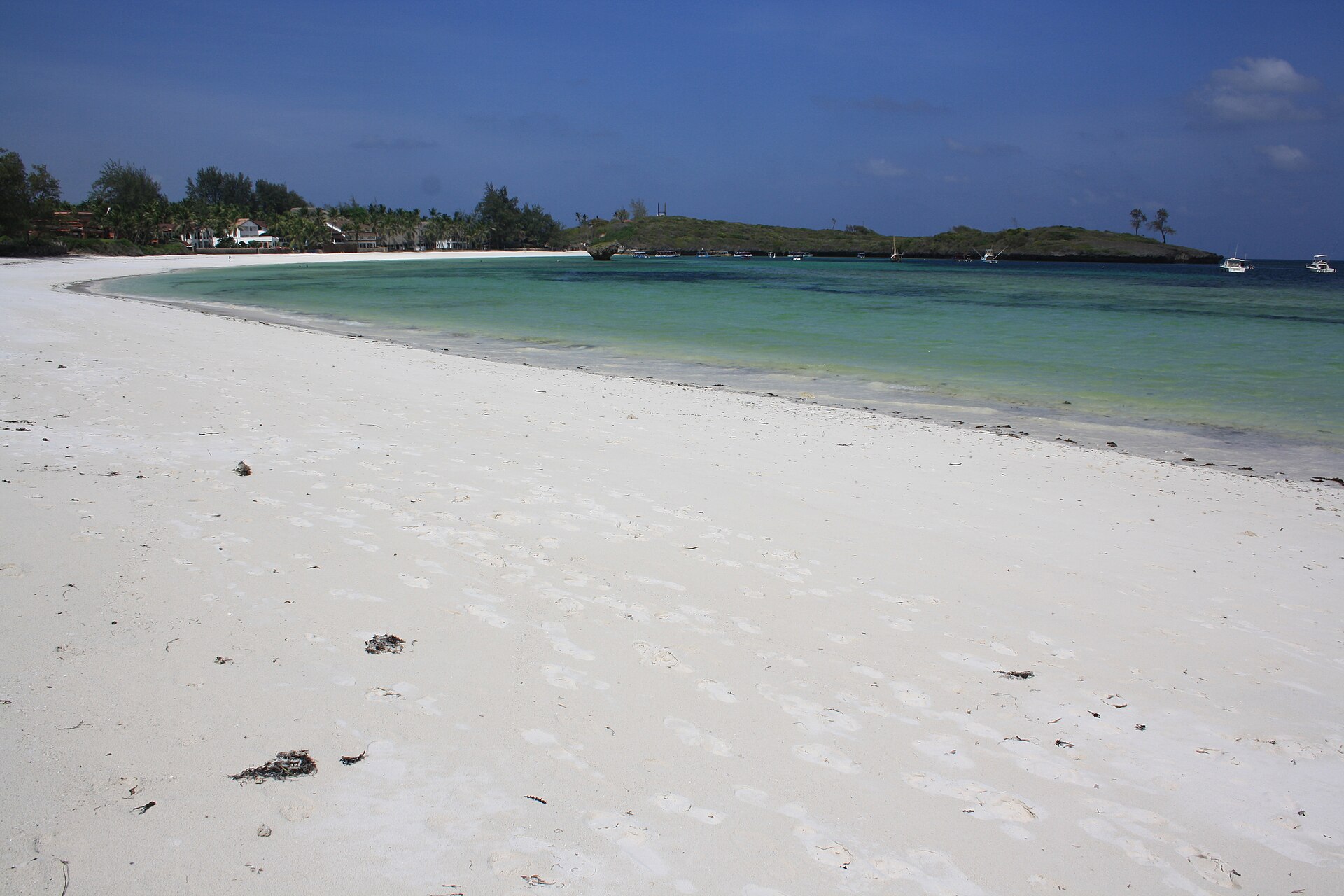 Weißer Sandstrand in weitem Bogen, türkises Wasser, dahinter eine bewachsene Landzunge und kleine Boote