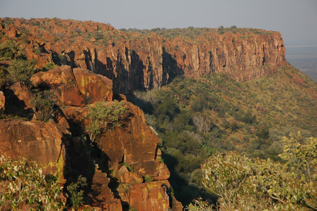 Blick vom Rand der Hochfläche auf die roten Sandsteinwände des Waterberg-Plateaus, darunter bewachsener Hang und Ebene