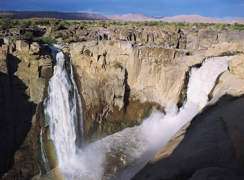 Zwei Wasserfälle stürzen in eine Granitschlucht, rechts der breite Hauptfall mit Gischt, links ein schmaler Seitenfall mit Regenbogen.