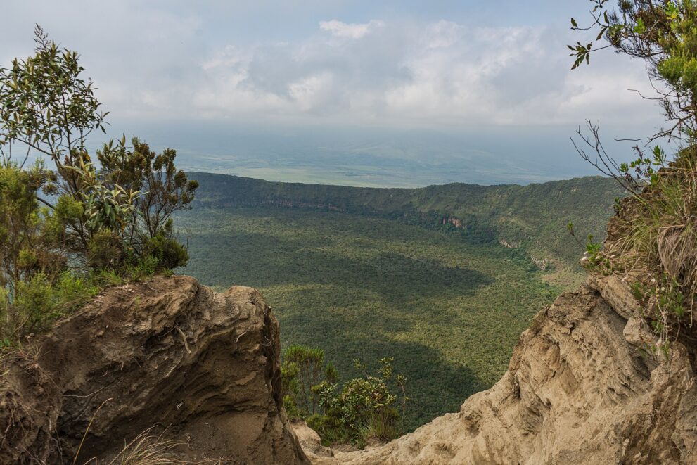 Zwischen zwei Felsen aus hellem Vulkangestein geht der Blick über den bewaldeten Krater hinweg in die dunstige Ebene des Rift Valley