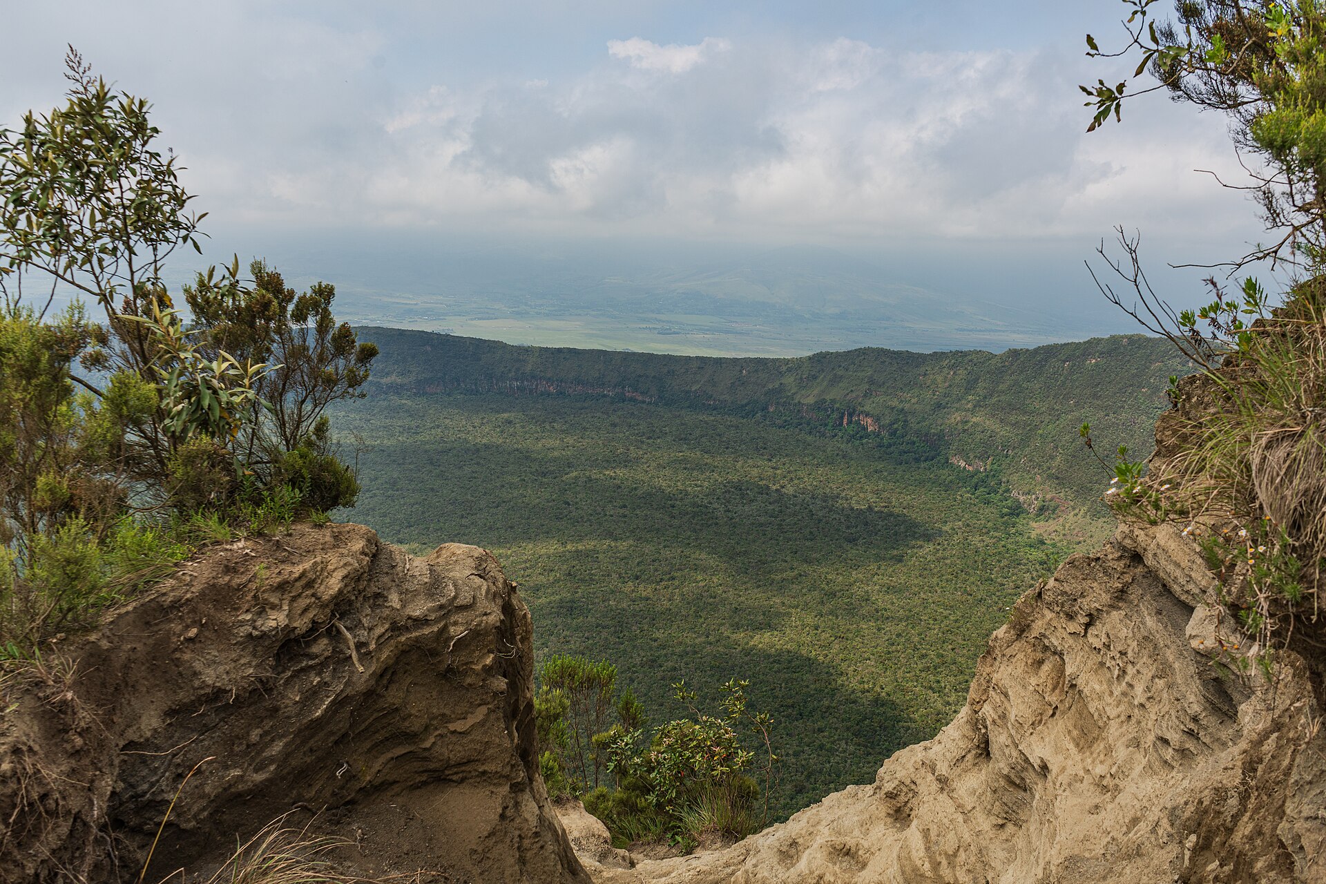 Zwischen zwei Felsen aus hellem Vulkangestein geht der Blick über den bewaldeten Krater hinweg in die dunstige Ebene des Rift Valley