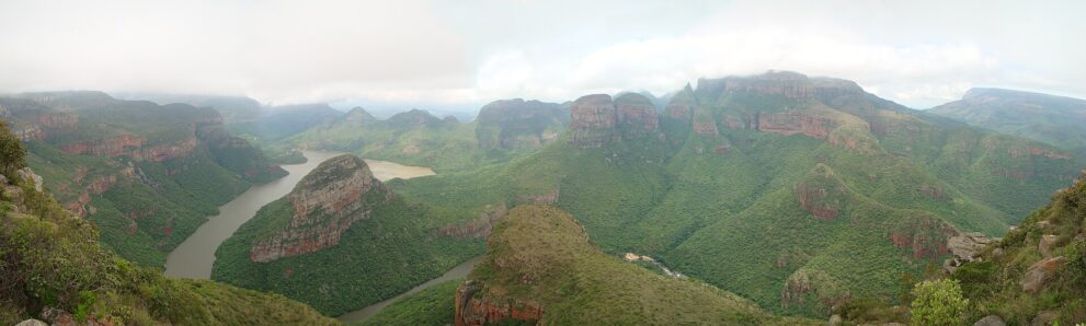 Weites Panorama über den grünen Canyon: links der Stausee zwischen Felsrücken, rechts die Three Rondavels unter tiefen Wolken.