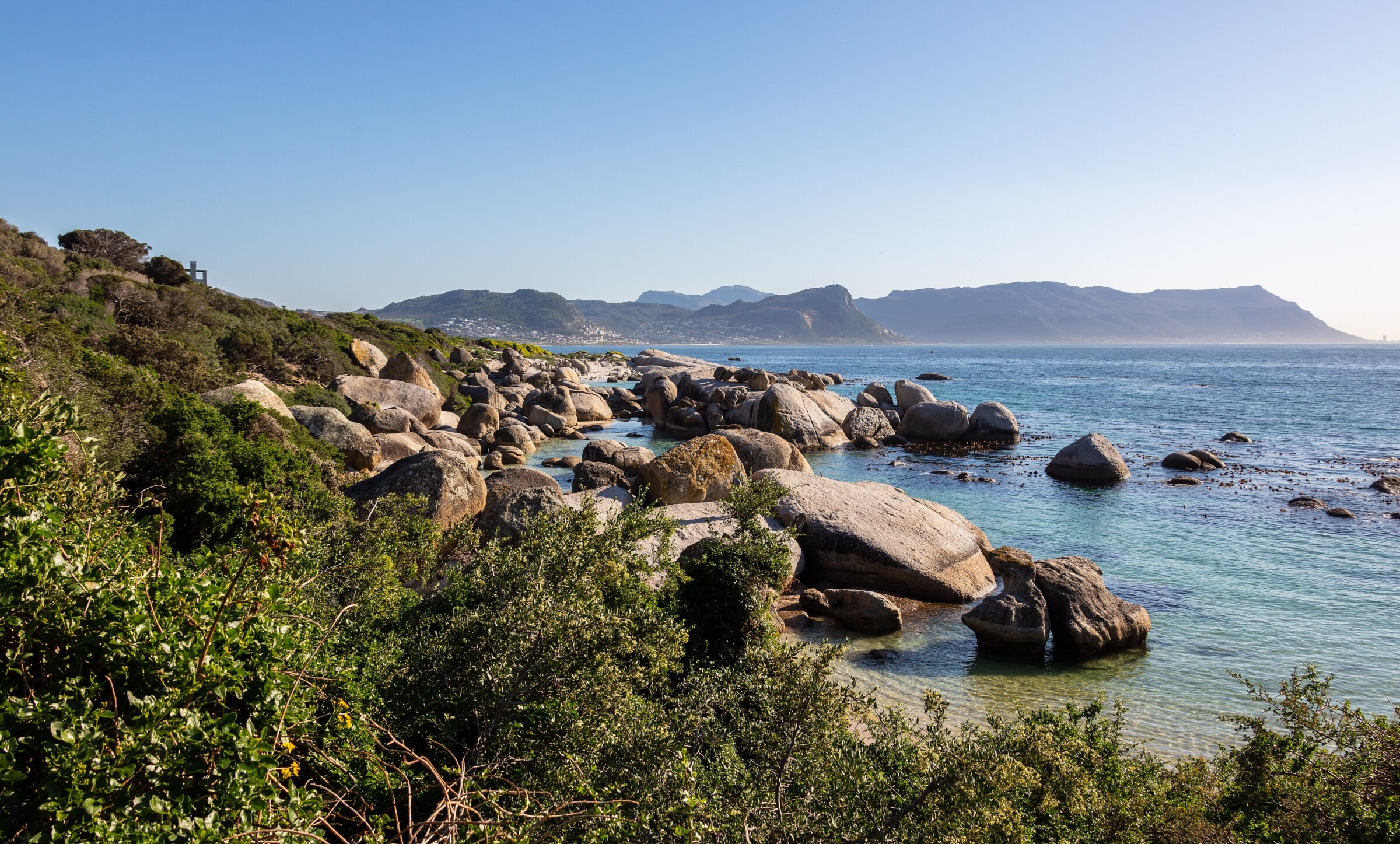 Runde Granitblöcke ziehen sich ins klare, flache Wasser, dahinter Berge am Ufer der False Bay