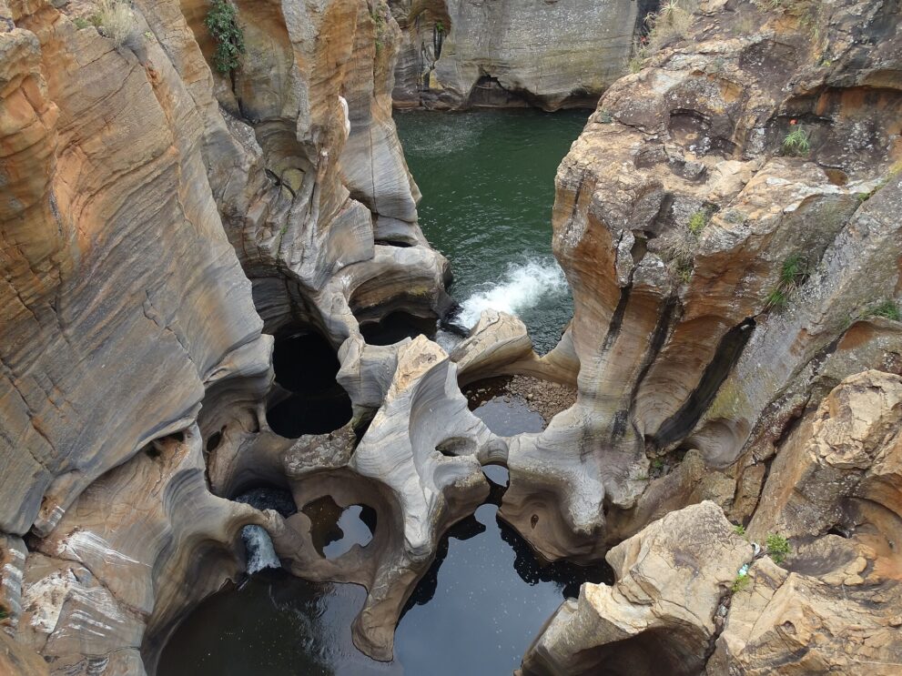Senkrechter Blick hinab auf hell geschliffene Felsbögen mit kreisrunden, dunklen Wasserlöchern, dahinter ein grünes Becken mit einer kleinen Kaskade