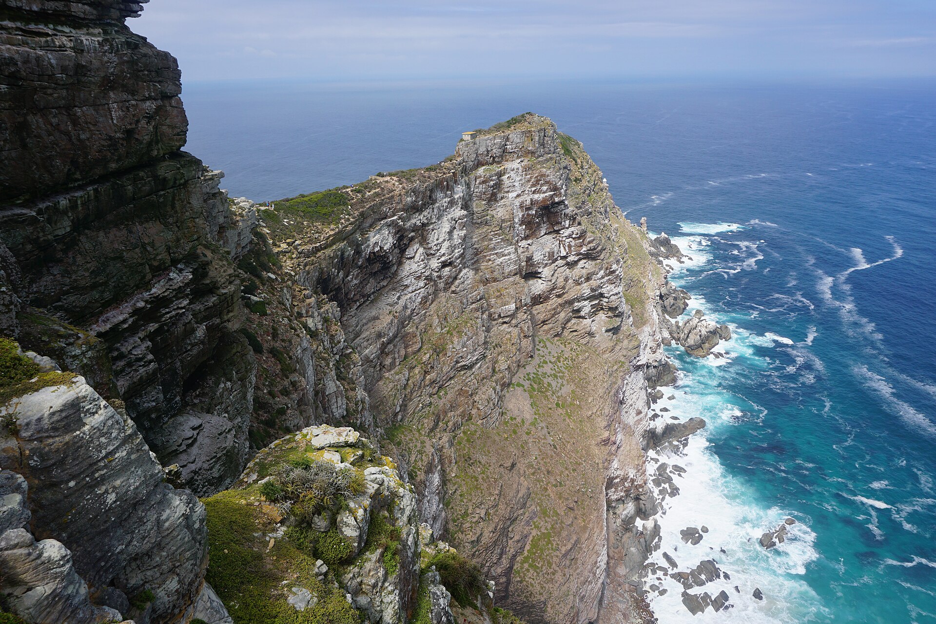 Steile, geschichtete Felsklippe, die als schmaler Grat ins tiefblaue Meer ragt, unten weiße Brandung an Felsen
